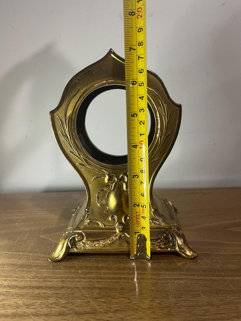 May include: A gold-coloured metal clock stand with an ornate design. The stand is approximately 15 cm tall.