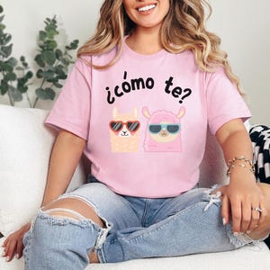 May include: A pink t-shirt with a graphic of two llamas wearing sunglasses and the text "¡cómo te?" printed on the front.