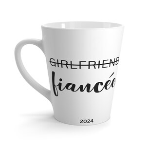 Może przedstawiać: Biały ceramiczny kubek latte z czarnym tekstem, który brzmi "GIRLFRIEND" przekreślone i "fiancée" napisane kursywą. Rok 2024 jest nadrukowany na spodzie kubka.