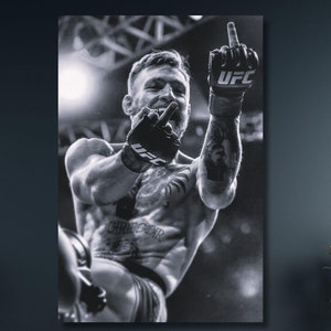 Peut inclure: Photographie en noir et blanc d'un homme dans un ring de boxe, levant le poing en l'air. Il porte des gants de boxe avec le mot "UFC" dessus.