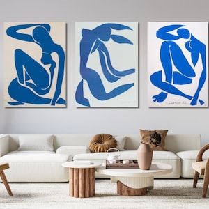 Può includere: Tre dipinti astratti blu di figure su sfondo bianco. I dipinti sono incorniciati e appesi a una parete grigio chiaro sopra un divano bianco, un tavolino da caffè e due sedie.