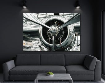 May include: A large rectangular wall art print featuring a close-up view of a polished silver vintage airplane engine with a prominent three-blade propeller. The metallic surface reflects light, highlighting the intricate mechanical details.