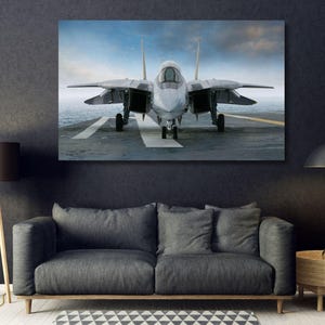 May include: A rectangular wall art print featuring a front-facing gray fighter jet parked on an aircraft carrier deck. The aircraft has dual vertical stabilizers, swept-back wings, and a cockpit canopy, set against a cloudy blue sky background.