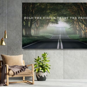 Op de afbeelding: Een rechthoekige wandkunstprint met een donkere, mistige weg omzoomd door dichte bomen, met de witte tekst HOLD THE VISION. TRUST THE PROCESS. in het midden bovenaan. De scène toont een verhard pad dat naar een mistige, lichte horizon leidt.