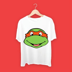 May include: White t-shirt with a green and orange Teenage Mutant Ninja Turtles character face graphic.