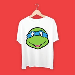 May include: White t-shirt with a green and blue cartoon turtle head graphic. The turtle is wearing a blue mask and has a big smile.
