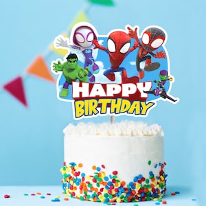 May include: A white cake with colorful sprinkles and a cake topper featuring Spider-Man, Spider-Gwen, Hulk, and Green Goblin characters. The topper says "Happy Birthday".