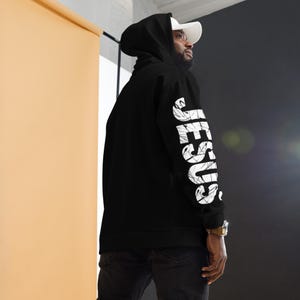 Puede incluir: Una sudadera negra con capucha que presenta la palabra JESUS impresa en letras mayúsculas blancas grandes con una textura marmoleada y desgastada que recorre verticalmente la parte exterior de la manga derecha. Tiene capucha con cordón.