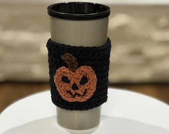 May include: A light gray tumbler with a black rim and a black crocheted sleeve. The sleeve features a centered, orange, hand-crocheted jack-o'-lantern pumpkin appliqué with a brown stem, dark eye cutouts, and a jagged mouth design.
