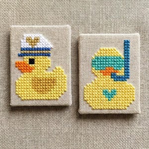 Könnte beinhalten: Zwei kleine, quadratische Kreuzstich-Kunstwerke. Eines zeigt eine gelbe Ente mit Matrosenmütze, blauem Band und goldenem Herz. Das andere zeigt eine gelbe Ente mit Schnorchel und Maske. Beide haben einen beigen Hintergrund.