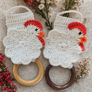 May include: Two crochet chicken-shaped towel holders featuring white yarn bodies, red combs, and orange beaks. Each chicken is attached to a circular wooden ring, with one ring in a light natural wood tone and the other in a dark brown wood finish.