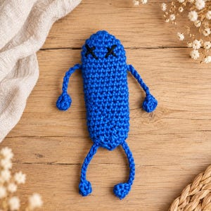 Könnte beinhalten: Eine kleine, leuchtend blaue Häkelfigur mit einem langen, abgerundeten Körper und dünnen, herabhängenden Armen und Beinen. Das Gesicht zeigt zwei schwarze, aufgestickte X-Formen als Augen, die einen einfachen Ausdruck auf der Garnoberfläche bilden.