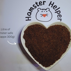 May include: A white heart-shaped bowl filled with 1 litre (approximately 300 grams) of brown hamster-safe soil. The bowl is on a white surface with the text "Hamster Helper" in a black circle with a cartoon hamster inside.