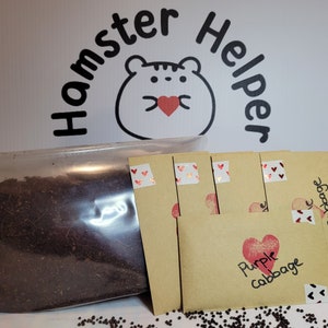 May include: A clear plastic bag filled with dark brown soil. Four brown paper envelopes with red hearts and the text "Purple Cabbage" are on a white surface. The envelopes are sealed with red heart stickers.