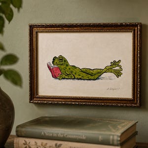 May include: A framed illustration of a green frog with dark spots lying on its stomach and reading a small red book. The artwork is presented in a rectangular, ornate, metallic gold frame with a textured border and a cream-colored background.