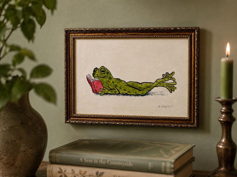May include: A framed illustration of a green frog with dark spots lying on its stomach and reading a small red book. The artwork is presented in a rectangular, ornate, metallic gold frame with a textured border and a cream-colored background.