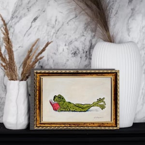 May include: A framed rectangular art print featuring a green frog lying on its stomach and reading a small red book. The illustration is set against a plain off-white background and is enclosed in a decorative, ornate gold-toned frame.