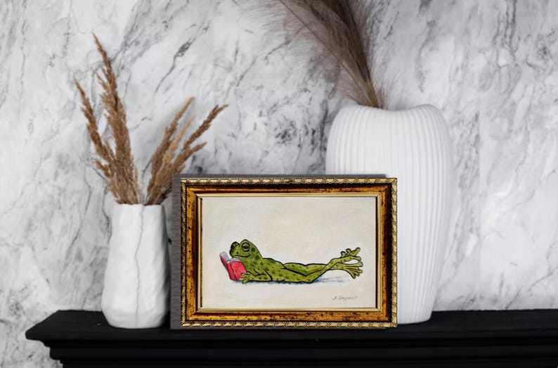 May include: A framed rectangular art print featuring a green frog lying on its stomach and reading a small red book. The illustration is set against a plain off-white background and is enclosed in a decorative, ornate gold-toned frame.