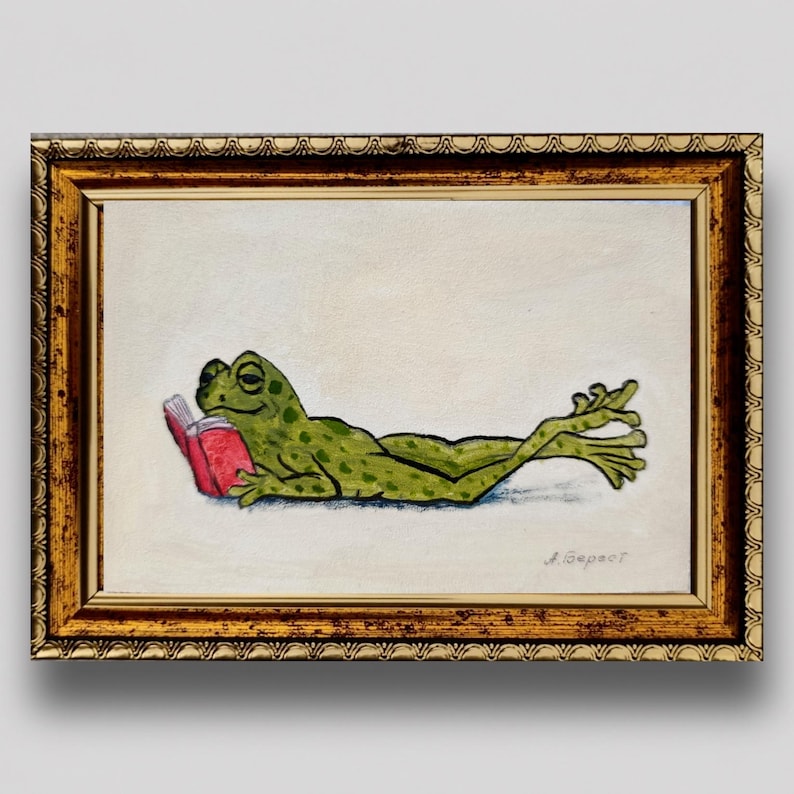 May include: A framed painting of a green frog with dark spots lying on its stomach while reading a small red book. The artwork is enclosed in an ornate, metallic gold-toned frame with a textured, scalloped edge and a signature in the corner.