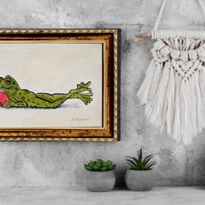 May include: A framed painting of a green frog with dark spots lying on its stomach and reading a small red book. The painting is enclosed in a decorative gold and black frame, featuring a signature in the bottom right corner of the artwork.