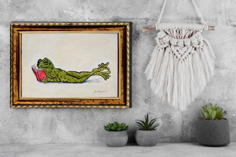 May include: A framed painting of a green frog with dark spots lying on its stomach and reading a small red book. The painting is enclosed in a decorative gold and black frame, featuring a signature in the bottom right corner of the artwork.