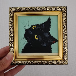 May include: A square painting of a black cat with bright yellow eyes set against a light green background. The cat is looking upward. The artwork is encased in an ornate, metallic gold-colored frame with intricate floral relief patterns.