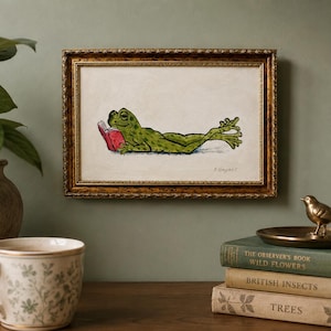 May include: A framed rectangular wall art piece featuring a green frog lying on its stomach and reading a small red book. The artwork is enclosed in an ornate, textured gold-colored frame with a cream-colored background and a signature in the corner.