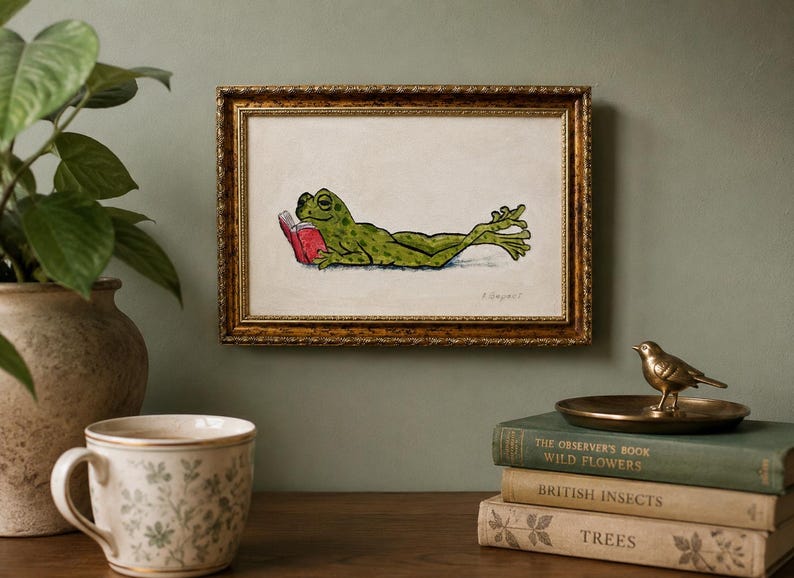 May include: A framed rectangular wall art piece featuring a green frog lying on its stomach and reading a small red book. The artwork is enclosed in an ornate, textured gold-colored frame with a cream-colored background and a signature in the corner.