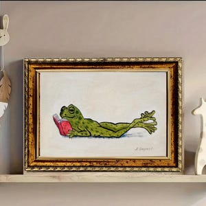 May include: A framed painting featuring a green frog with dark spots lying on its stomach while reading a small red book. The artwork is set against a light cream background and is enclosed in a decorative, textured gold-toned rectangular frame.