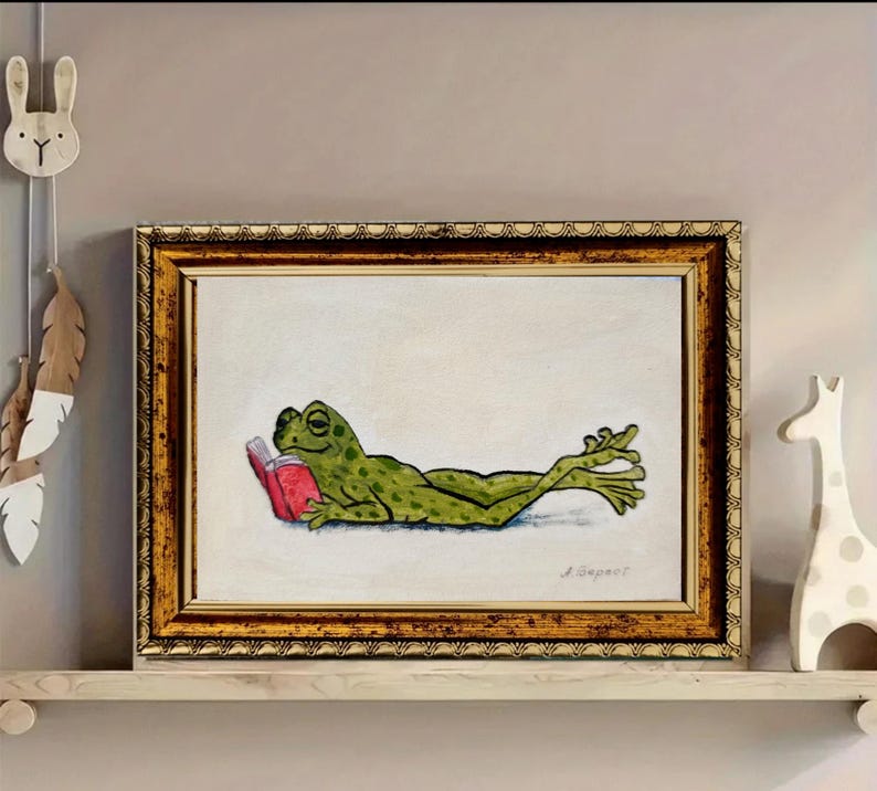 May include: A framed painting featuring a green frog with dark spots lying on its stomach while reading a small red book. The artwork is set against a light cream background and is enclosed in a decorative, textured gold-toned rectangular frame.