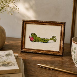May include: A rectangular picture frame with an ornate gold border containing a watercolor illustration of a green frog lying down and reading a small red book. The frog is depicted in a relaxed pose with its legs extended, set against a plain background.