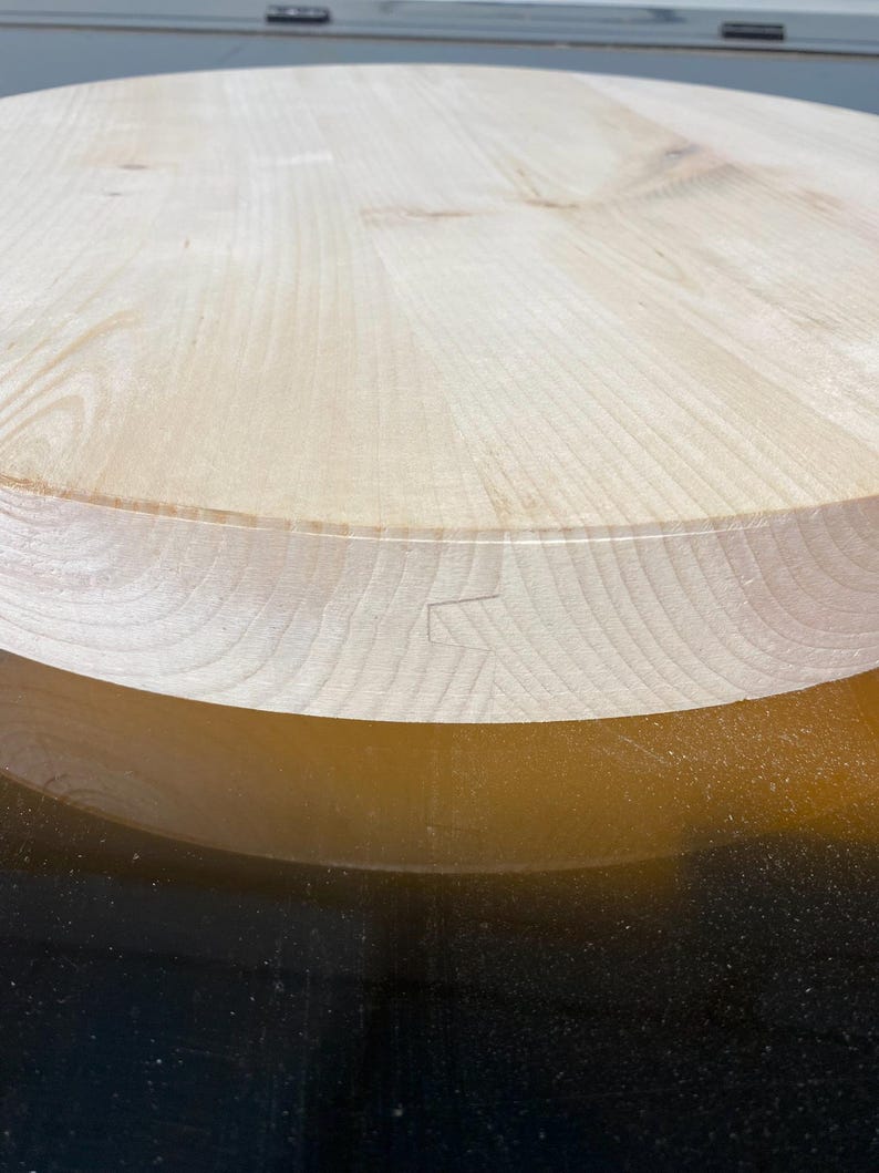 May include: Close-up of a large, unfinished, round wooden tabletop. The light-coloured wood grain is visible, with a detailed edge and a visible joint. The table is likely for a dining or kitchen area.