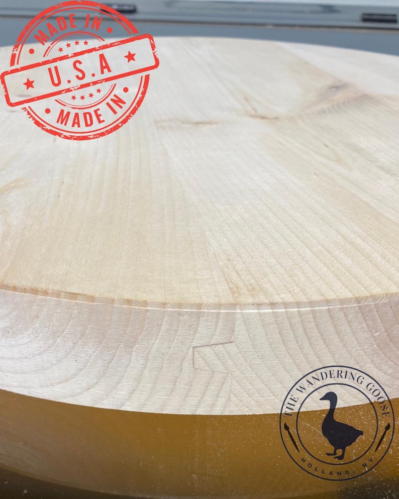 May include: Close-up of a round, light-coloured wooden surface. The wood grain is visible. A red stamp with the text "MADE IN U.S.A." is at the top. The bottom right corner has a logo with a goose silhouette.
