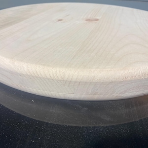 May include: A light-coloured, round wooden tabletop with a smooth, unfinished surface. The wood grain is visible, and the edges are slightly rounded. The tabletop is likely for a table or similar furniture.