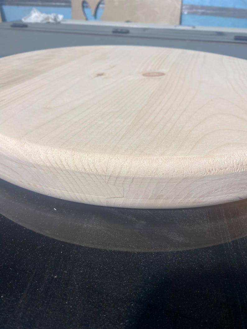 May include: A light-coloured, round wooden tabletop with a smooth, unfinished surface. The wood grain is visible, and the edges are slightly rounded. The tabletop is likely for a table or similar furniture.