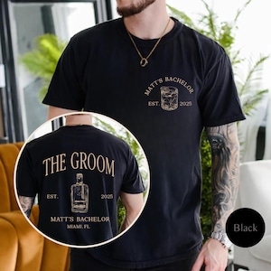 May include: Black t-shirt with gold text and graphics. The front features a whiskey glass design with "MATT'S BACHELOR EST. 2025". The back displays "THE GROOM" above a liquor bottle graphic, "EST. 2025" and "MATT'S BACHELOR MIAMI. FL".