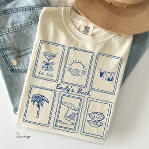 May include: An ivory t-shirt featuring six blue rectangular stamp-style graphics. The designs include a martini glass with 'Est. 2026', a sunset, a clothesline with swimwear, a palm tree, a beach umbrella with 'Miami, FL', and a seashell. Text reads 'Emily's Bach'.