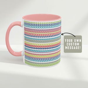 May include: A white ceramic mug with a pink interior and handle. The mug features a colorful, repeating pattern of horizontal lines in various colors. The text "YOUR OWN CUSTOM MESSAGE!" is on a tag attached to the mug.