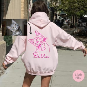 May include: Pale pink hoodie with a pink cat illustration and the name "Bella" in cursive. The hoodie has a drawstring hood and long sleeves. A small image of a cat is in the upper left corner. The text "Ligth Pink" is in a pink circle.