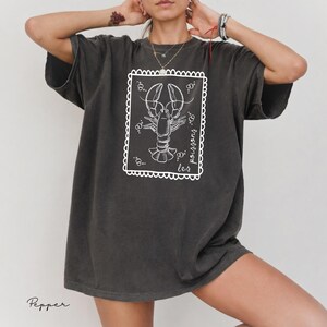 May include: Dark gray t-shirt featuring a white illustration of a lobster inside a decorative frame. The text "les poissons" is written in cursive on the right side of the frame. The shirt has short sleeves and a relaxed fit.