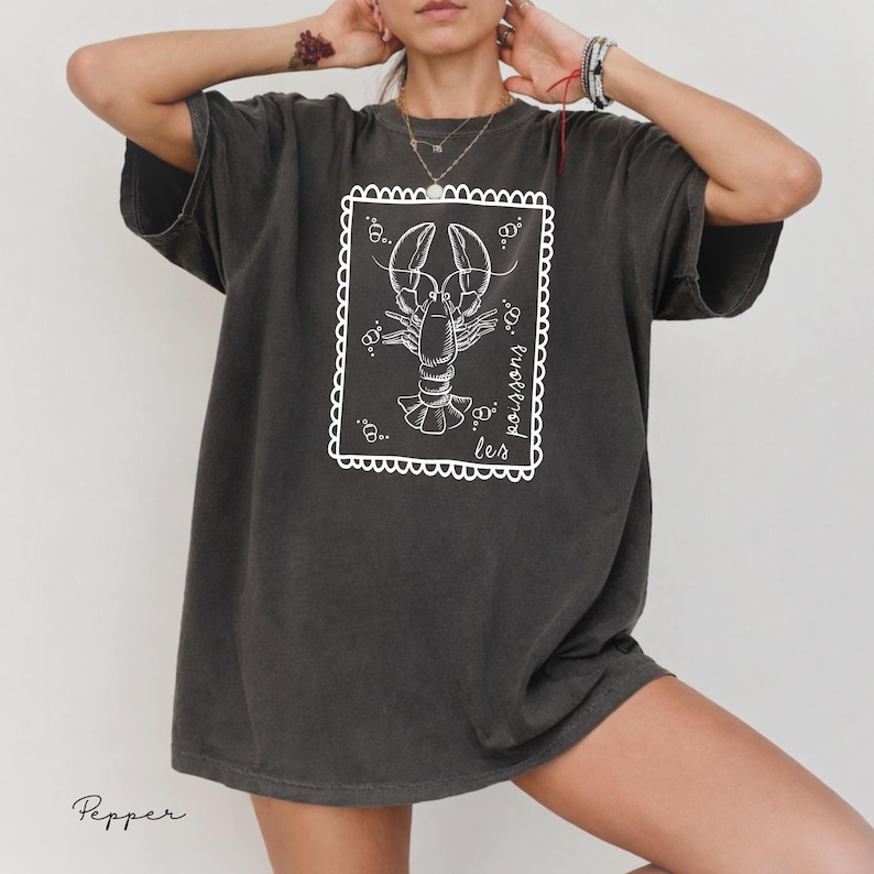 May include: Dark gray t-shirt featuring a white illustration of a lobster inside a decorative frame. The text "les poissons" is written in cursive on the right side of the frame. The shirt has short sleeves and a relaxed fit.