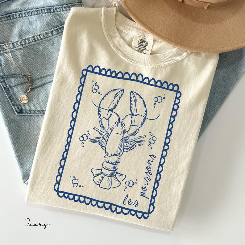 May include: Ivory-colored t-shirt featuring a blue lobster illustration within a scalloped border. The text "les poissons" is written in blue. The shirt is made of Comfort Colors material.