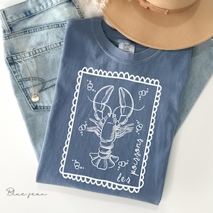 May include: A blue t-shirt with a white lobster illustration inside a decorative frame. The shirt is laid on top of blue jeans, with a gold necklace and a tan hat in the background. The text "les poissons" is printed on the shirt.