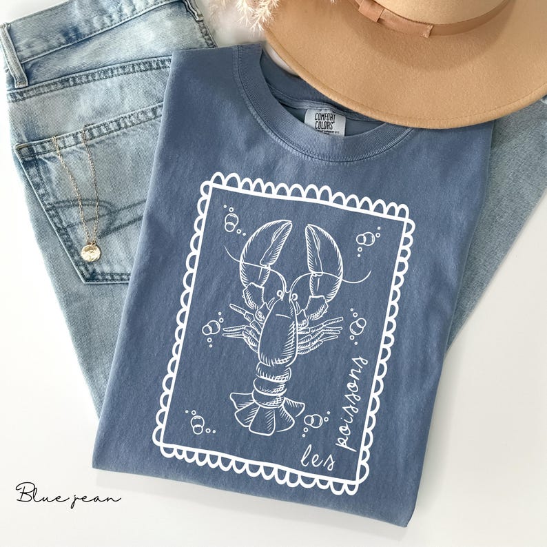 May include: A blue t-shirt with a white lobster illustration inside a decorative frame. The shirt is laid on top of blue jeans, with a gold necklace and a tan hat in the background. The text "les poissons" is printed on the shirt.