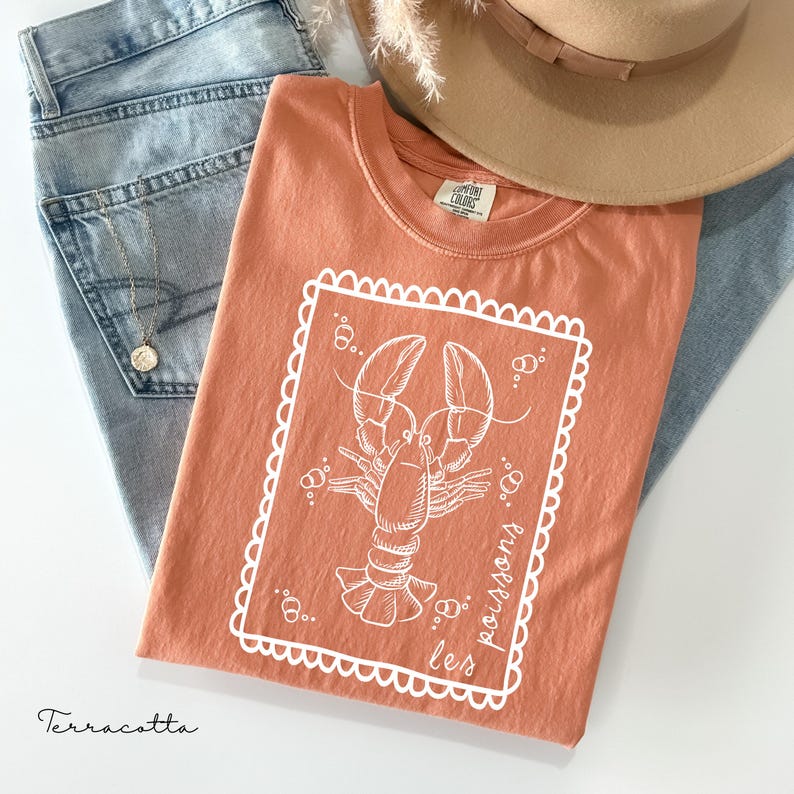 May include: Terracotta-colored t-shirt with a white lobster graphic inside a scalloped frame. The shirt is folded on top of blue jeans, with a gold necklace and a tan hat in the background. The text "les poissons" is printed on the shirt.