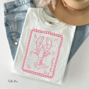 May include: White t-shirt with a pink lobster graphic inside a pink scalloped frame. The text "les poissons" is printed in pink on the right side of the frame. The shirt is folded on top of blue jeans, with a tan hat and a necklace visible.