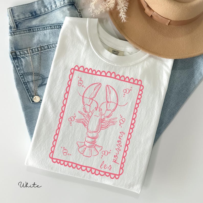 May include: White t-shirt with a pink lobster graphic inside a pink scalloped frame. The text "les poissons" is printed in pink on the right side of the frame. The shirt is folded on top of blue jeans, with a tan hat and a necklace visible.