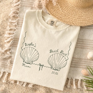 May include: Cream-colored t-shirt with a seashell design and the text "Kaylee's Beach Bach Miami 2026". The shirt is made of a soft material and is laying on a beach towel with a straw hat.