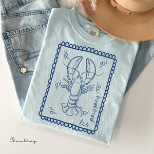 May include: Light blue t-shirt with a navy blue lobster graphic inside a scalloped border. The words "les poissons" are printed on the shirt. The word "Chambray" is printed at the bottom left.