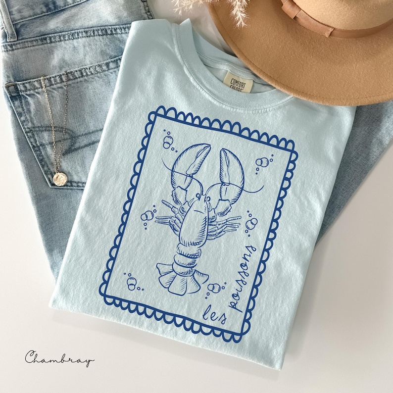 May include: Light blue t-shirt with a navy blue lobster graphic inside a scalloped border. The words "les poissons" are printed on the shirt. The word "Chambray" is printed at the bottom left.